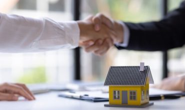 Two people shaking hands over a desk with a small model house in the foreground, symbolizing a home purchase agreement and the importance of securing home insurance before closing.