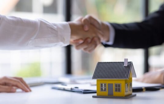 Two people shaking hands over a desk with a small model house in the foreground, symbolizing a home purchase agreement and the importance of securing home insurance before closing.