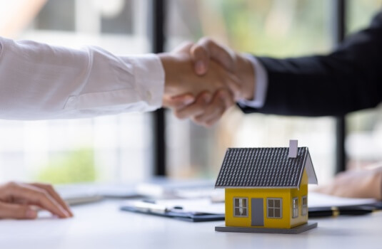 Two people shaking hands over a desk with a small model house in the foreground, symbolizing a home purchase agreement and the importance of securing home insurance before closing.