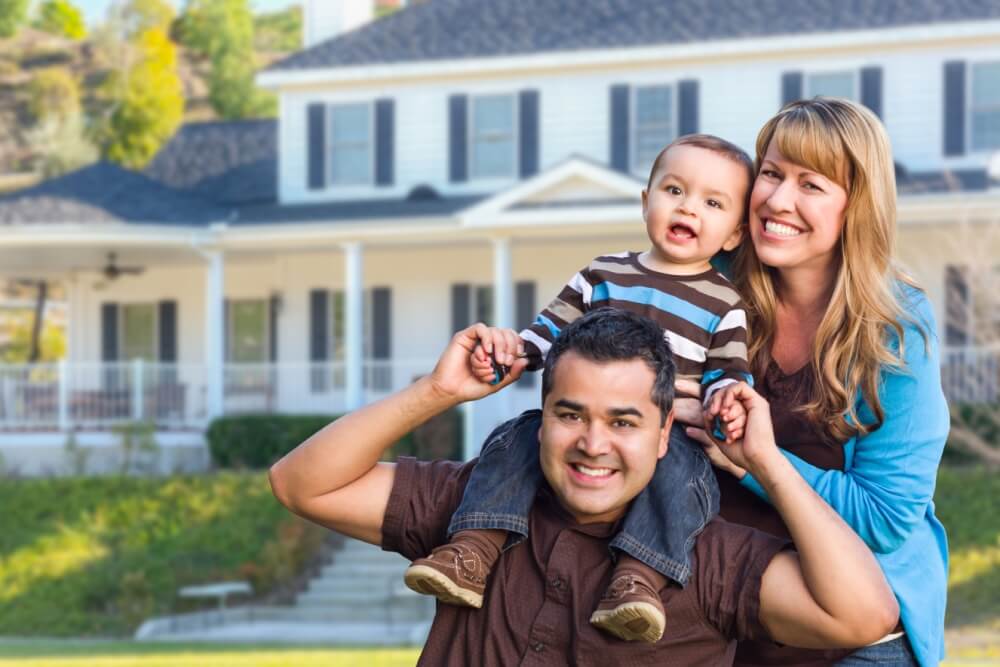 A smiling family standing in front of their home, with a father carrying a child on his shoulders and a mother beside them, highlighting the importance of home insurance for protecting your home and loved ones.