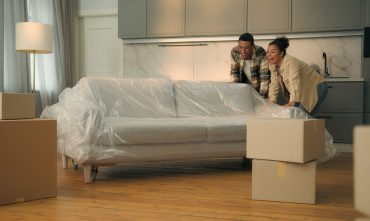 Happy couple moving a plastic-covered couch into their new apartment, surrounded by moving boxes — ideal image representing renters insurance, protecting personal belongings in a rental home.