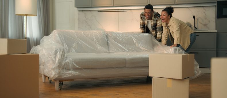 Happy couple moving a plastic-covered couch into their new apartment, surrounded by moving boxes — ideal image representing renters insurance, protecting personal belongings in a rental home.