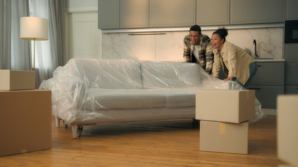 Happy couple moving a plastic-covered couch into their new apartment, surrounded by moving boxes — ideal image representing renters insurance, protecting personal belongings in a rental home.