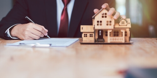 Close-up of a homeowner signing paperwork next to a model house, representing Georgia homeowners insurance policies, claims, and property coverage decisions.