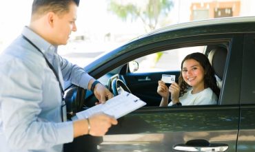 Young new driver sitting in a car smiles and holds up a driver's license while speaking with an insurance representative standing outside the vehicle and reviewing paperwork on a clipboard. The scene suggests obtaining or discussing auto insurance coverage for a new driver.