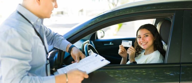 Young new driver sitting in a car smiles and holds up a driver's license while speaking with an insurance representative standing outside the vehicle and reviewing paperwork on a clipboard. The scene suggests obtaining or discussing auto insurance coverage for a new driver.