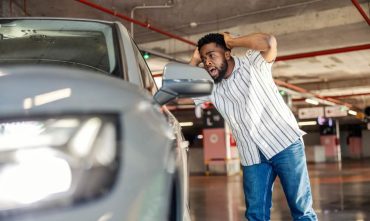 Man reacts with concern while inspecting damage on his car in a parking garage and wondering if auto insurance cover scratches.