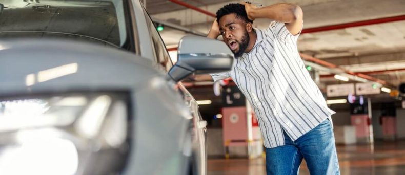Man reacts with concern while inspecting damage on his car in a parking garage and wondering if auto insurance cover scratches.