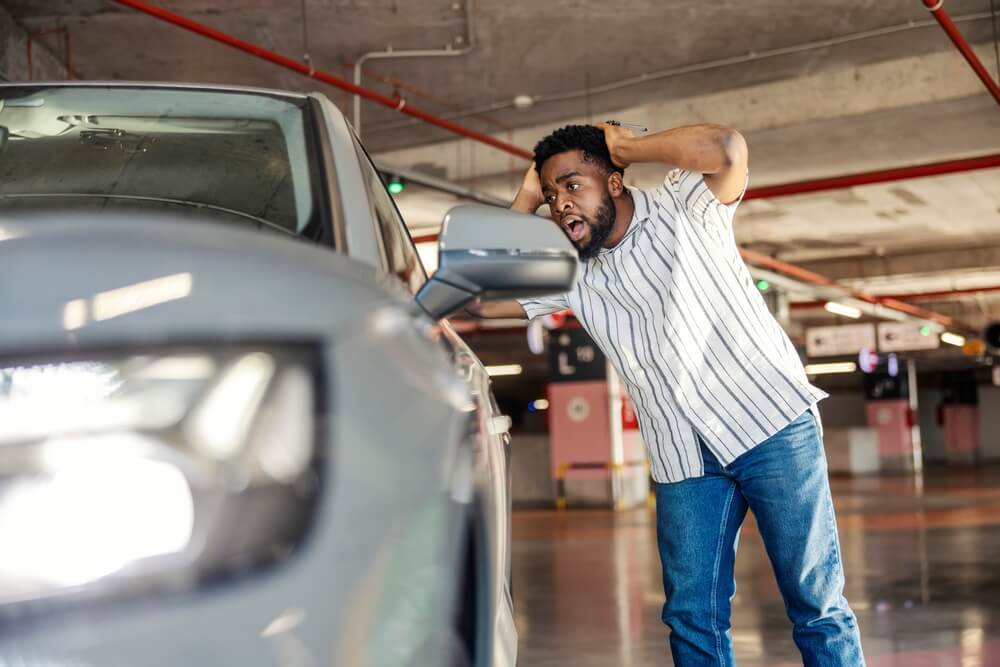 Man reacts with concern while inspecting damage on his car in a parking garage and wondering if auto insurance cover scratches.
