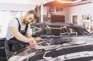 Detailer polishing a scratch on a dark car inside an auto shop while the owner considers whether auto insurance cover scratches.