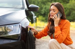 Woman kneels beside her car while checking a scratch near the front wheel and calling for help about whether auto insurance cover scratches.
