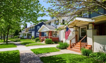 Row of townhouses with front porches, green lawns, and trees in a residential neighborhood.
