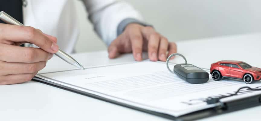 Hand with pen signing a paper with car keys and toy car to represent insurance policy