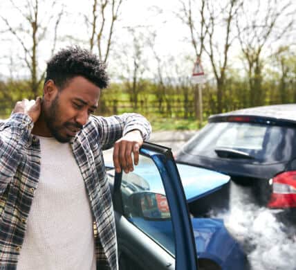 Worried african american young man holding his neck with a car accident in the background