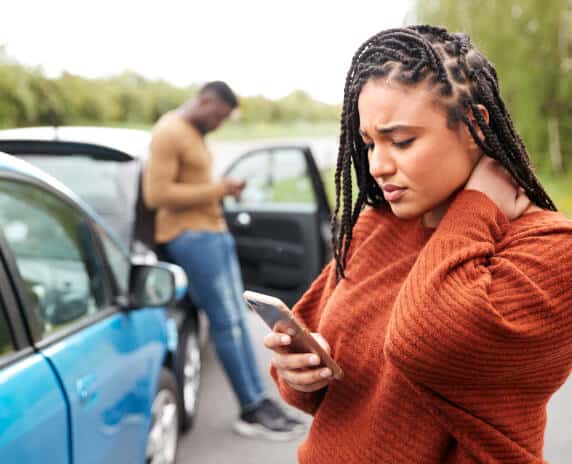 Worried african american woman looking at phone with a car accident in the background