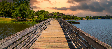 a brown wooden bridge over the silky green waters surrounded by lush green trees and plants with powerful clouds at sunset in Georgia.