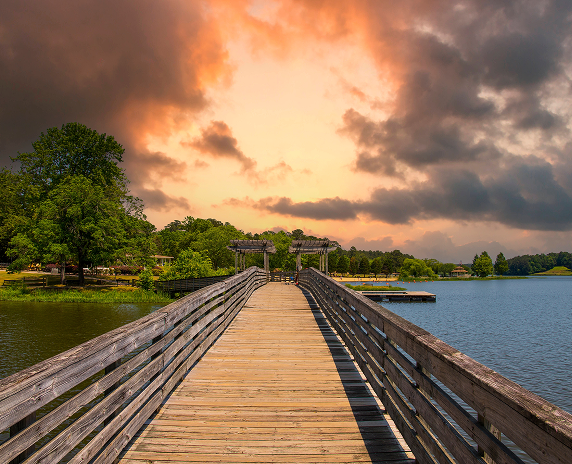 a brown wooden bridge over the silky green waters surrounded by lush green trees and plants with powerful clouds at sunset in Georgia.