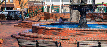 A circular red brick water fountain with benches, bare winter trees and a little girl running and parked cars at the Marietta Square in Marietta - cheap car insurance in Georgia.