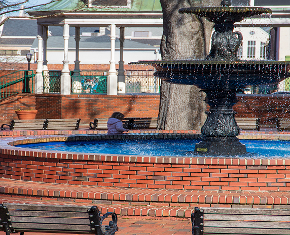 A circular red brick water fountain with benches, bare winter trees and a little girl running and parked cars at the Marietta Square in Marietta - cheap car insurance in Georgia.