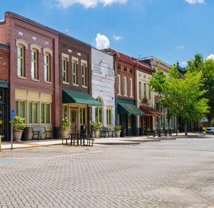 A sunny downtown street in Powder Springs, Georgia, lined with historic brick storefronts and awnings, with “City Hall” visible on one building, trees providing shade, and cars parked along a wide, paved main street. Cheap car insurance in Georgia.
